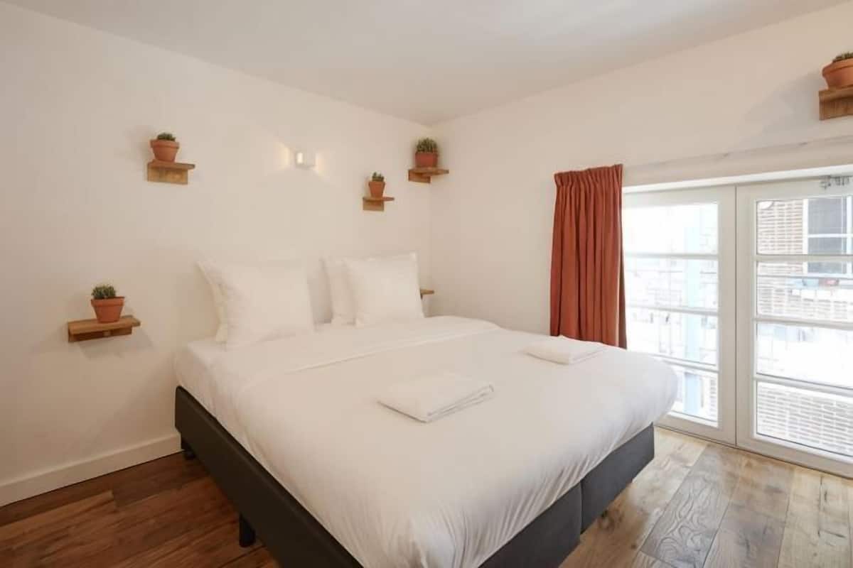 A bedroom with a bed, white bedding, wooden flooring, and potted plants on shelves.