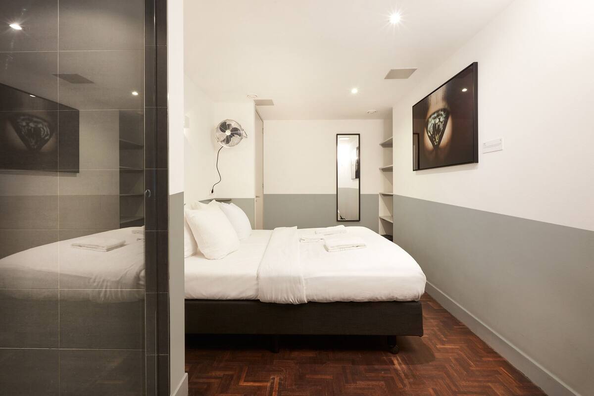 A hotel room with a bed, a mirror, a fan, and a painting on the wall.
