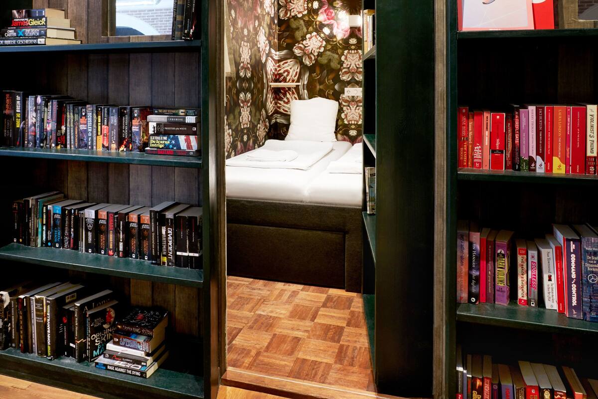 A hotel room with a bed, a bookshelf, and a window.