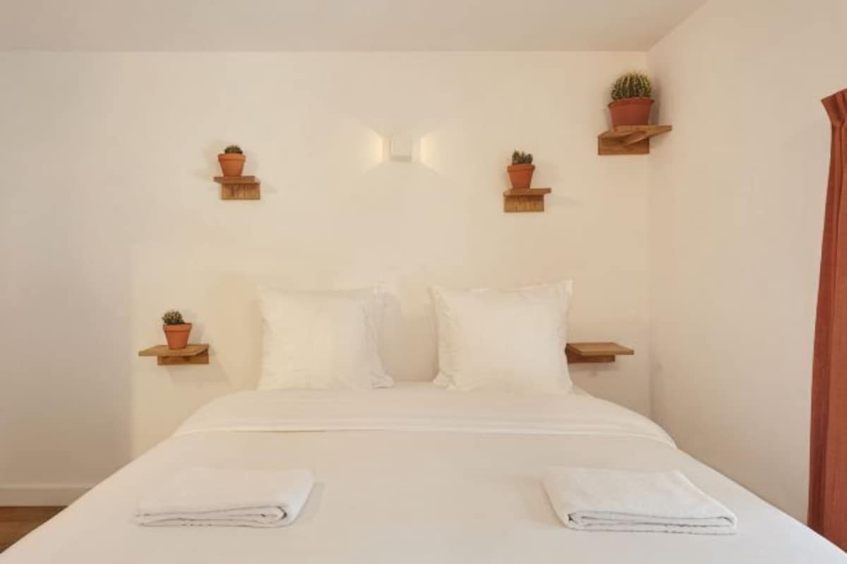 Una camera da letto con un letto, cuscini, una coperta ordinatamente ripiegata e piante in vaso sugli scaffali.