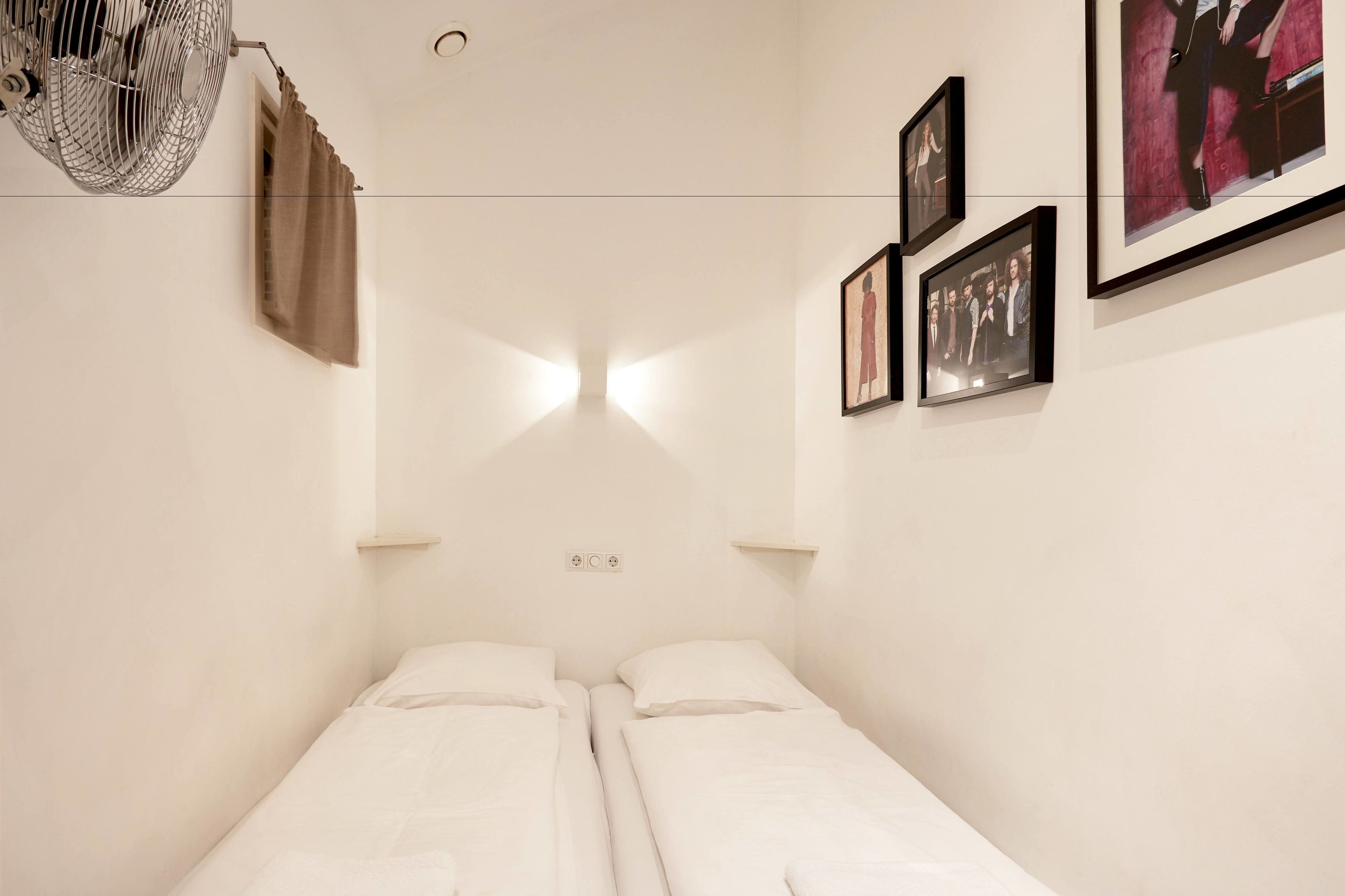 A minimalist room with two single beds, a ceiling fan, a wall-mounted shelf, and framed pictures.