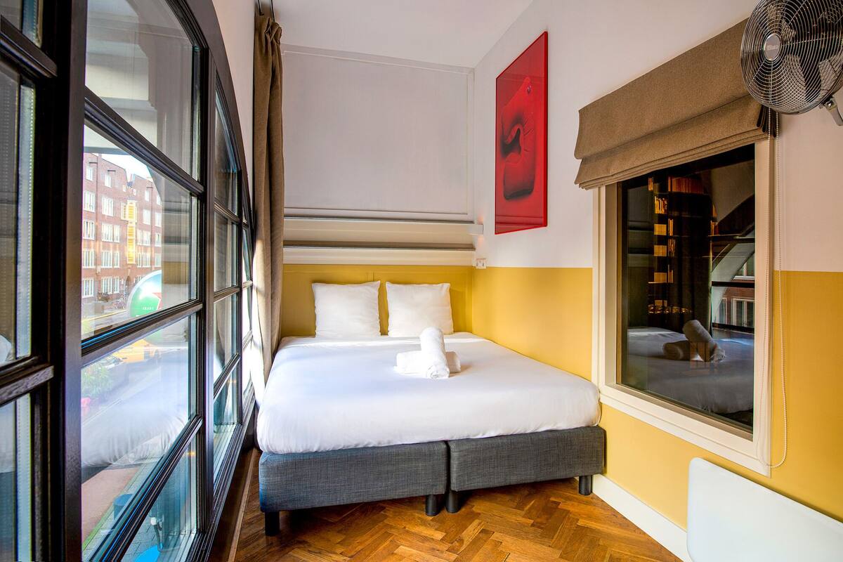 A hotel room with a large window, a bed with white linens, a grey bench, and a red artwork on the wall.