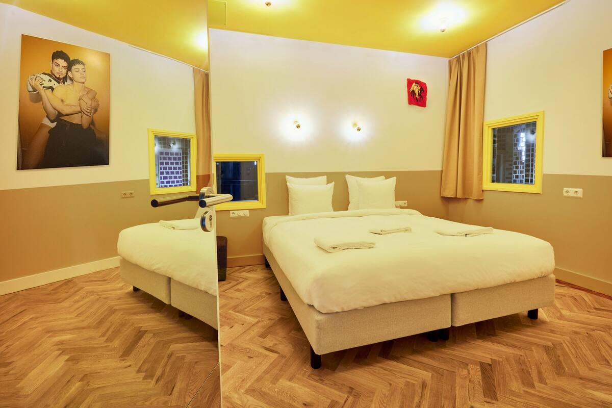 A hotel room with a large bed, a smaller bed, a window with curtains, a wall-mounted TV, and a painting on the wall.