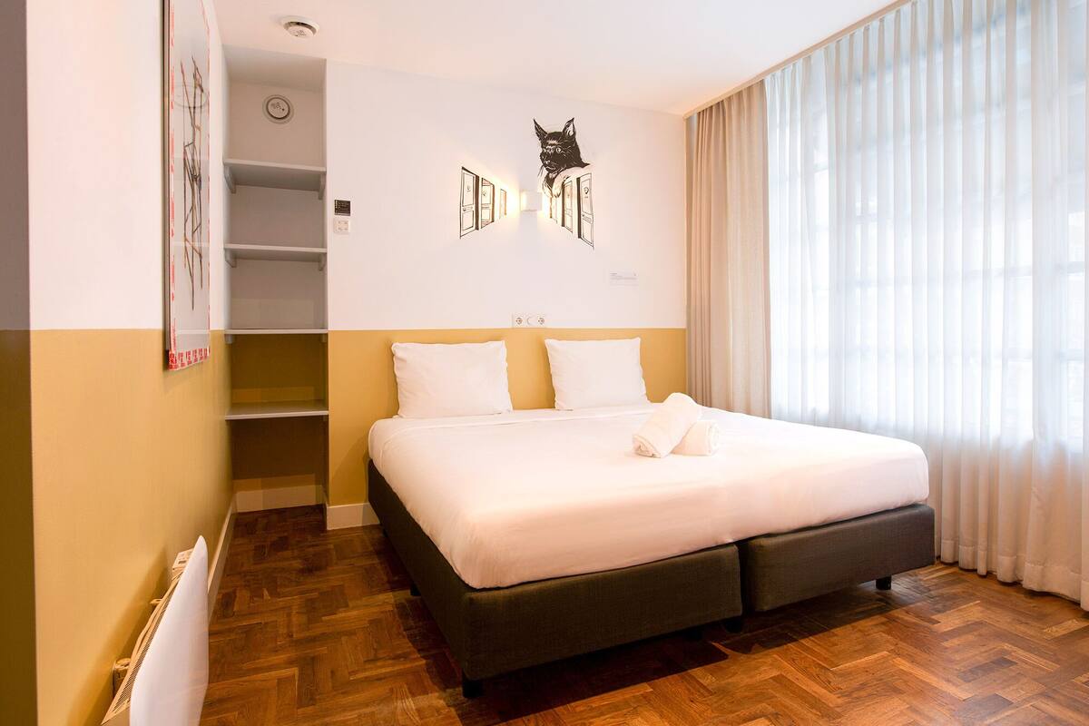 A hotel room with a large bed, a wardrobe, a cat wall art, and a window with curtains.