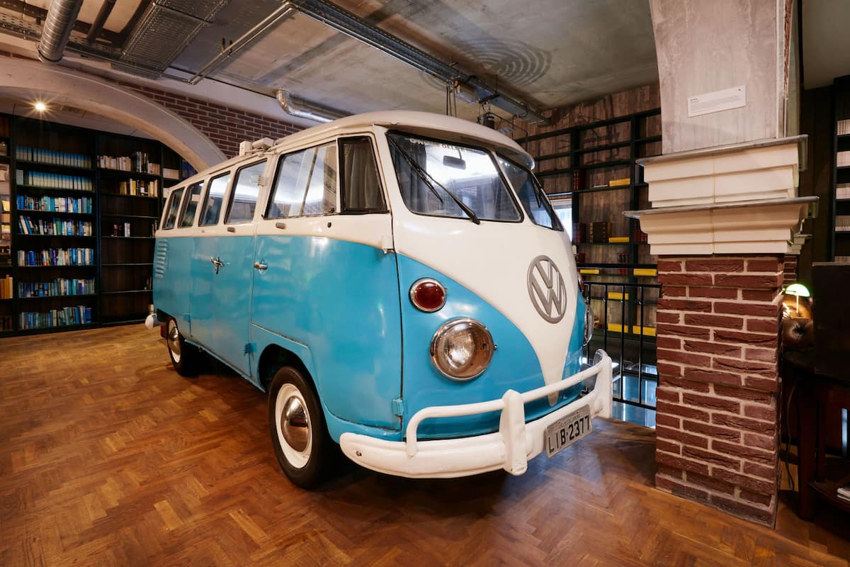 A vintage Volkswagen van in a library setting with bookshelves and a brick wall.