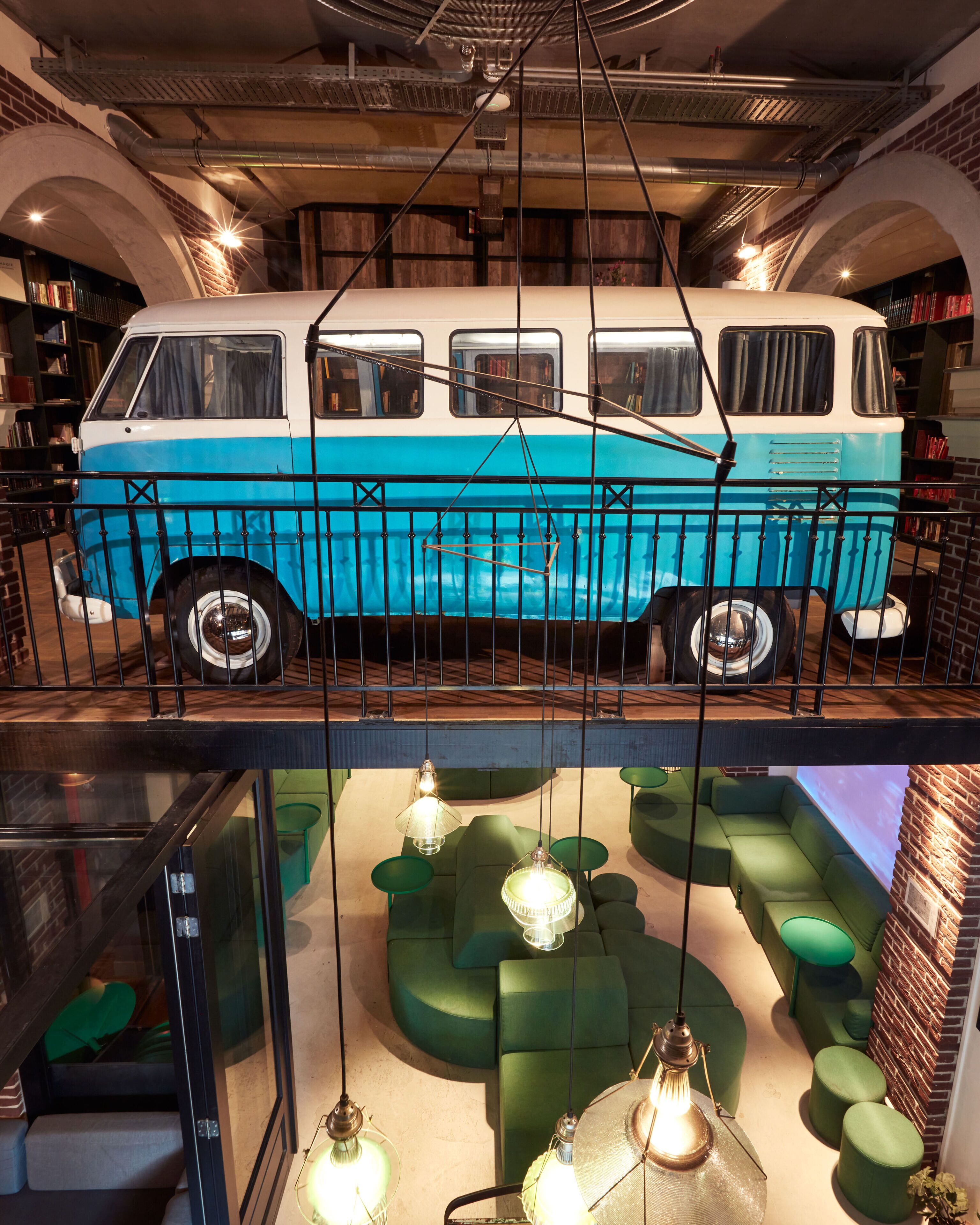 Un vieux fourgon bleu et blanc est exposé dans une pièce où se trouvent des canapés verts et des lampes suspendues.