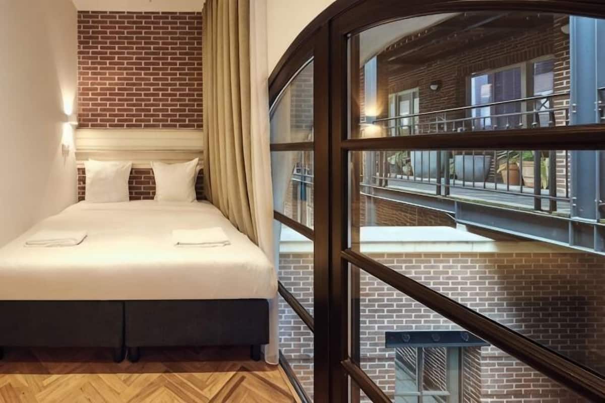 A hotel room with a bed, a brick wall, a window, and a view of the outside.