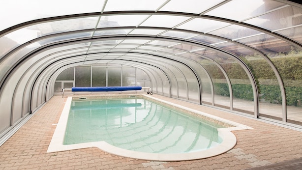 An indoor swimming pool with a glass ceiling, surrounded by brick flooring and a blue bench. - Hôtel Alpen Roc (Morzine)