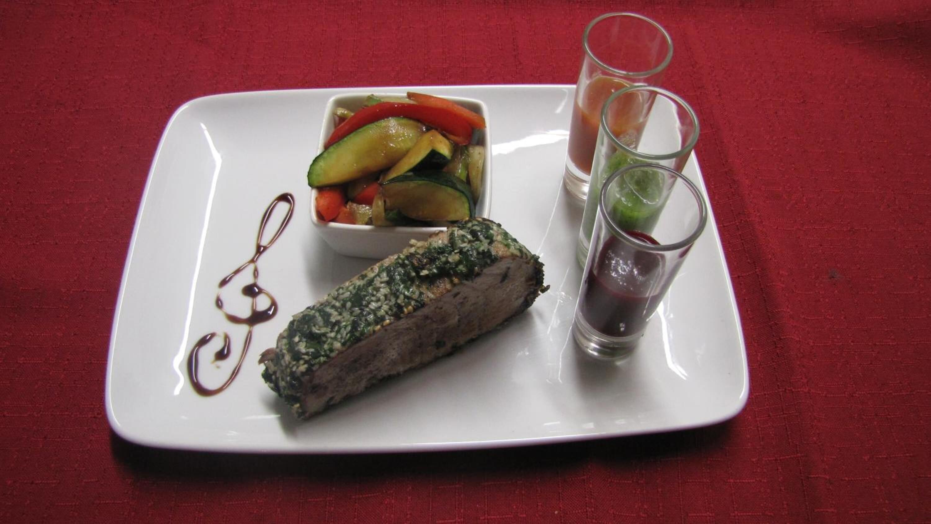 Un plato blanco con un trozo de carne cubierto de hierbas, un bol de verduras variadas y dos vasos de un líquido oscuro.