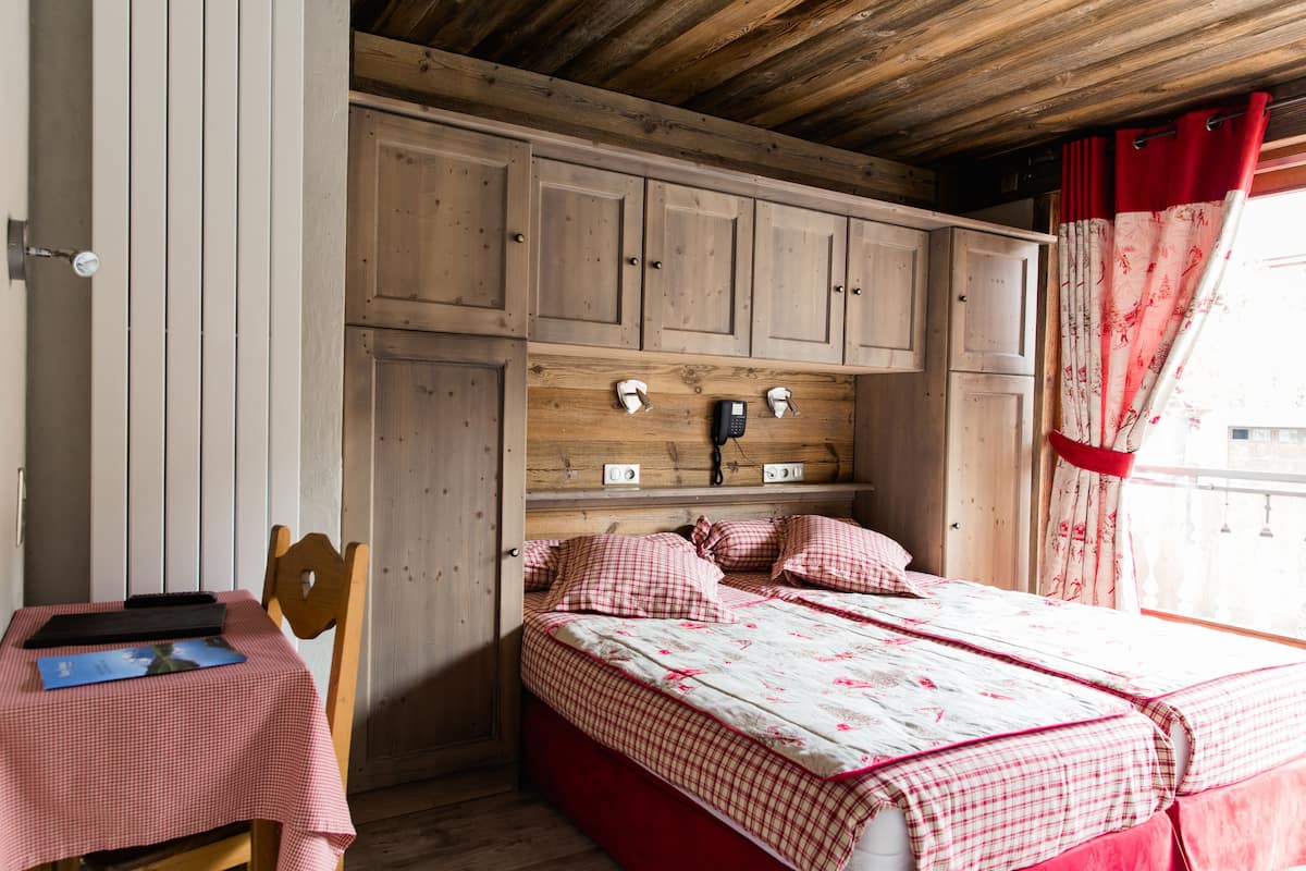 A bedroom with a wooden headboard, a bed with checkered bedding, a bedside table with a book and a phone, and a window with red curtains.