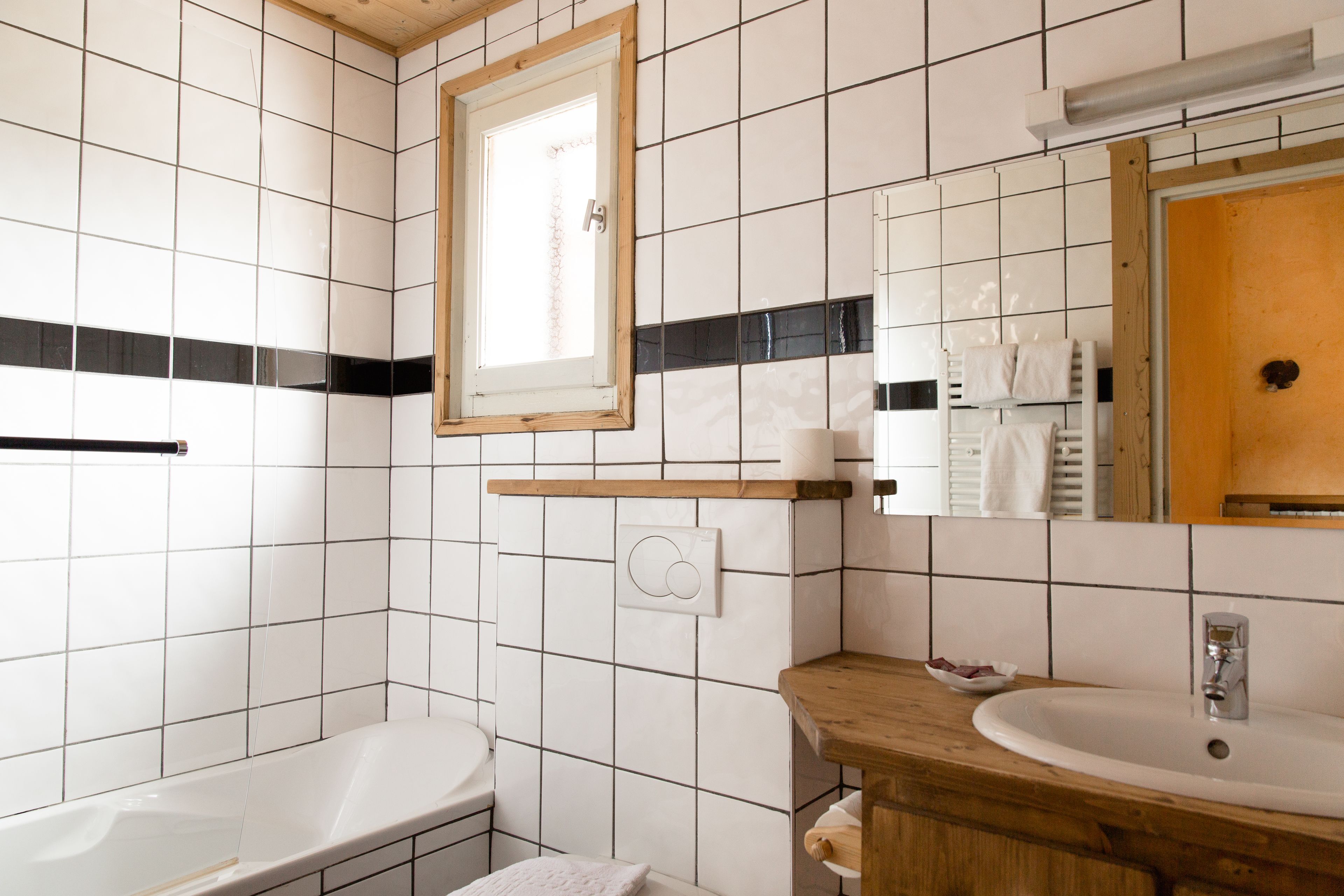 Een badkamer met witte betegelde wanden, een houten wastafelmeubel, een wastafel, een spiegel en een raam.