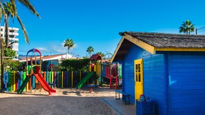 A colorful playground with slides and climbing structures, a small blue building with a yellow door, and palm trees in the background.