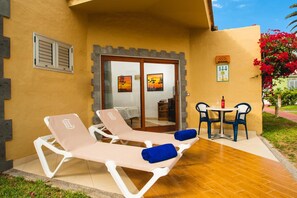 A sunlit outdoor area with two lounge chairs and a table, a door leading to an interior space, and a small garden with a red flowering plant. - HL Miraflor Suites (San Bartolomé de Tirajana)