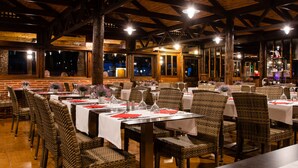 A restaurant with multiple tables and wicker chairs, wooden beams on the ceiling, and large windows.