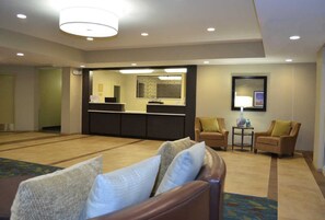 Un hall de hotel con mostrador de recepción, una zona de estar con dos sillones y una mesita, y un espejo grande en la pared.