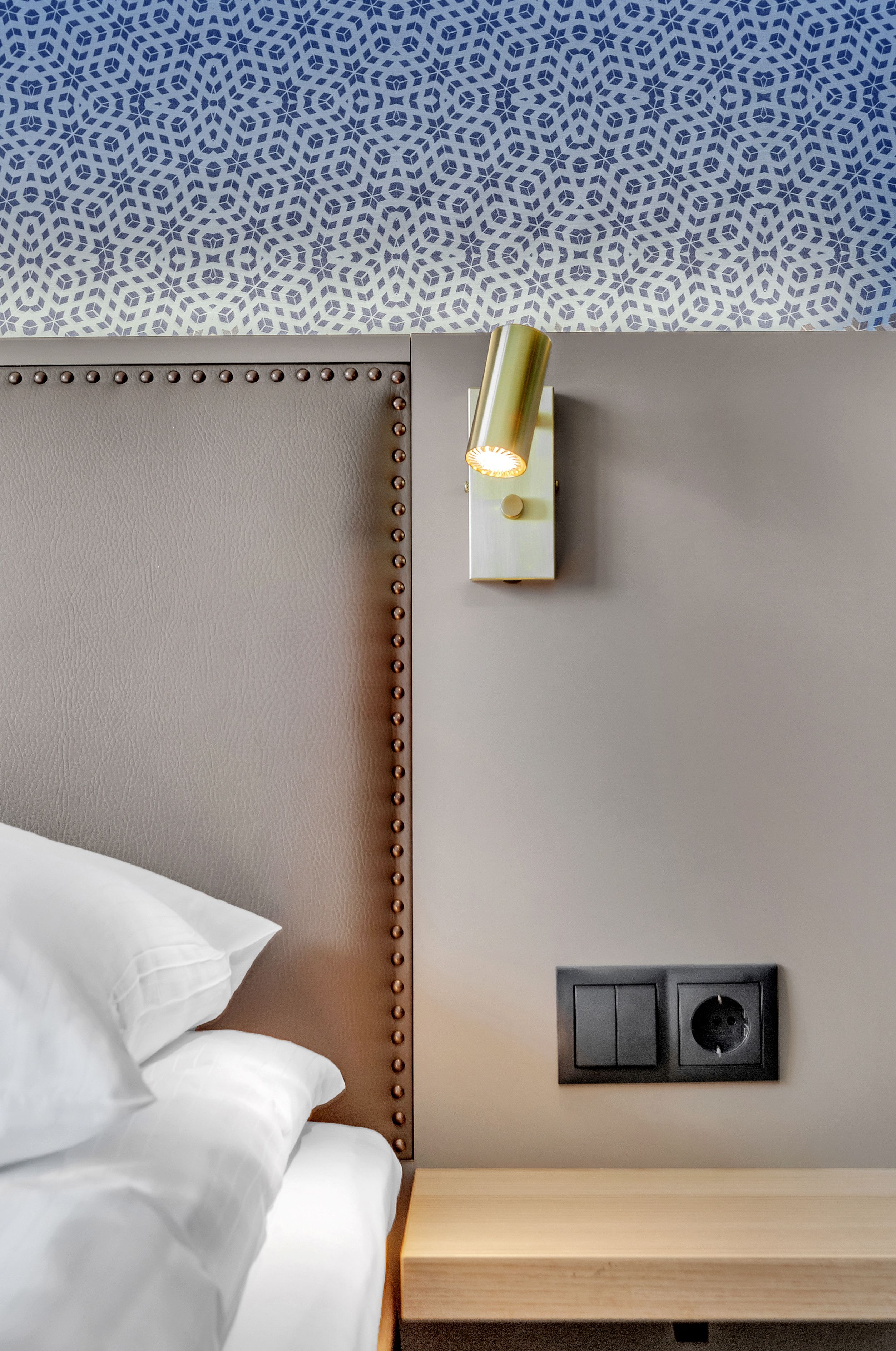 Una habitación de hotel con una cama, una mesita de noche, una lámpara de pared y un enchufe.