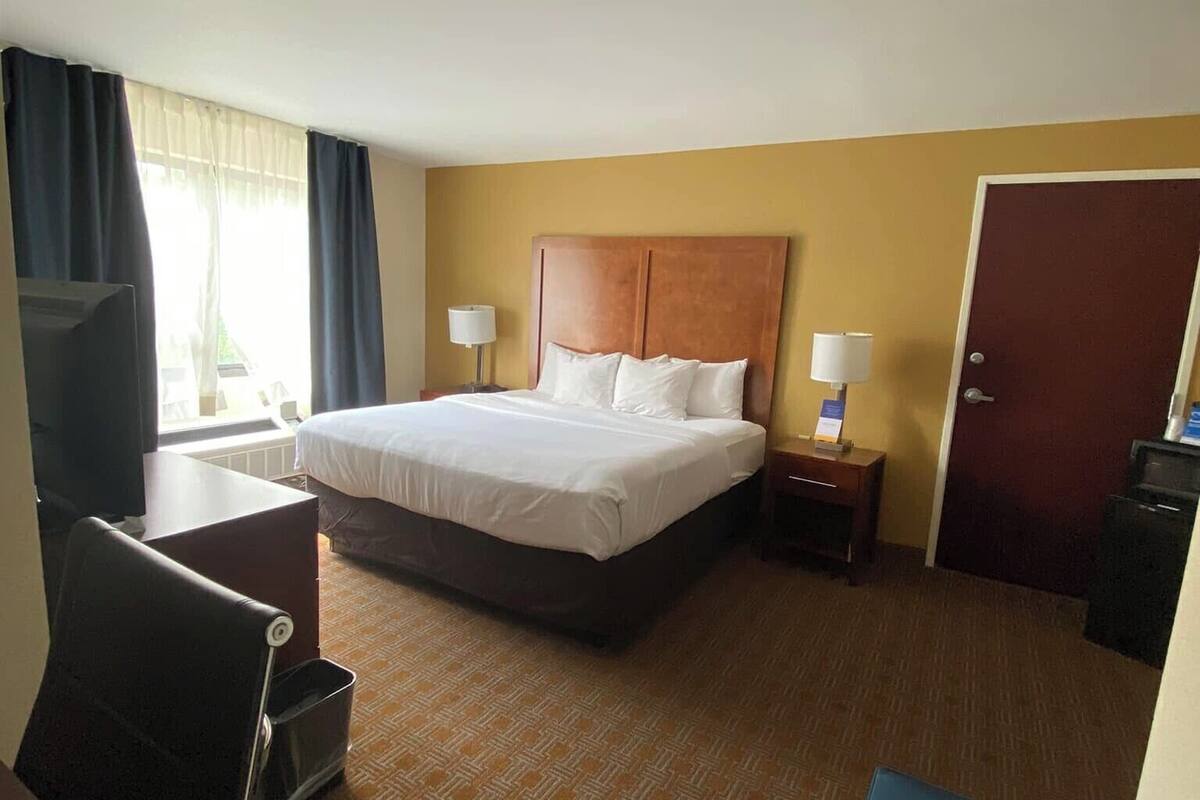 A hotel room with a large bed, two bedside tables with lamps, a desk with a chair, a TV, and a door.