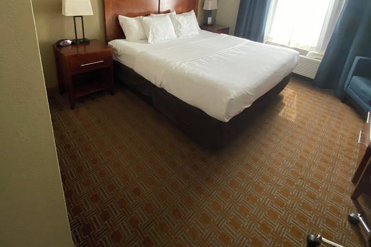 A hotel room with a large bed, a wooden headboard, a nightstand with a lamp, and a window with curtains.