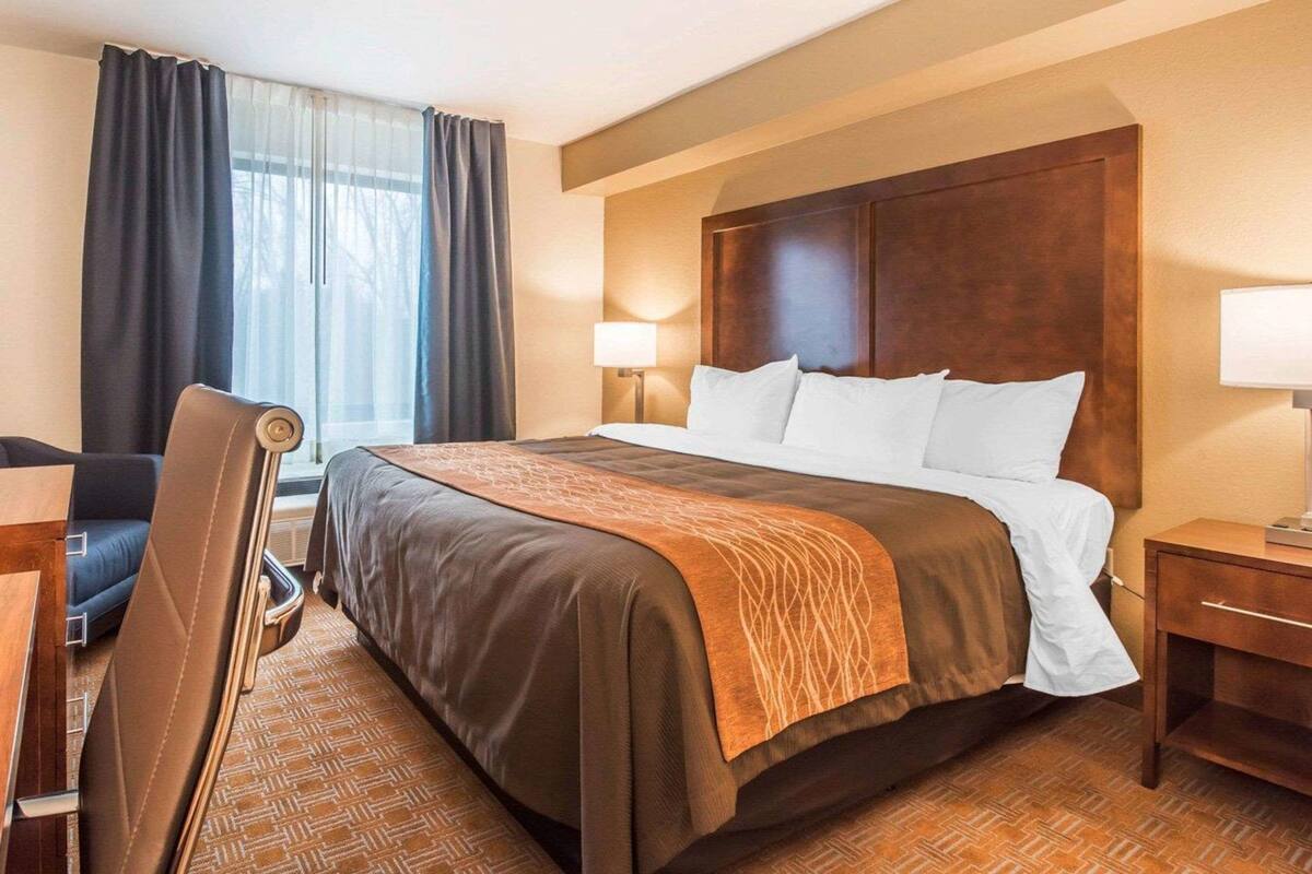 A hotel room with a large bed, a desk, a chair, a nightstand, and a window with curtains.