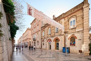 Um edifício histórico com uma grande faixa, área de pedestres com azulejos e uma loja com um letreiro vermelho em formato de 'A'.