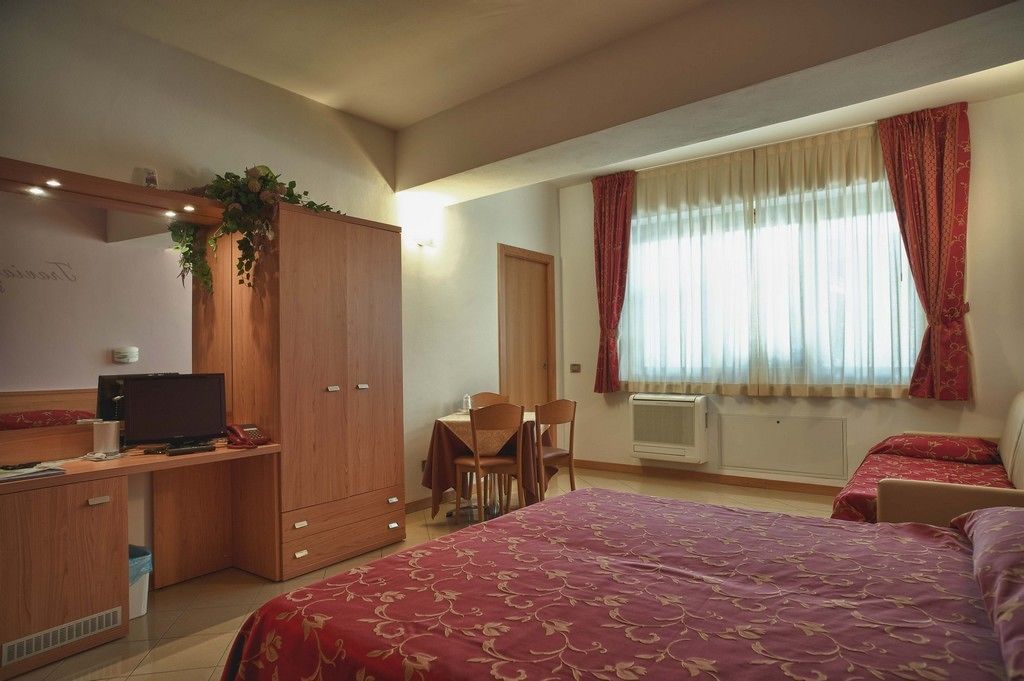 Hotellihuone, jossa on sänky, työpöytä tietokoneella, tuoli, vaatekaappi ja ikkuna, jossa on verhot.