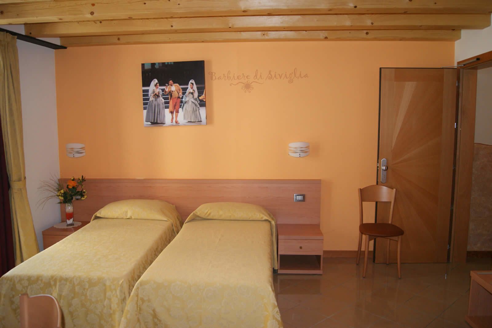 A hotel room with two beds, a wooden headboard, a wall-mounted picture, a nightstand with a vase of flowers, and a wooden chair.