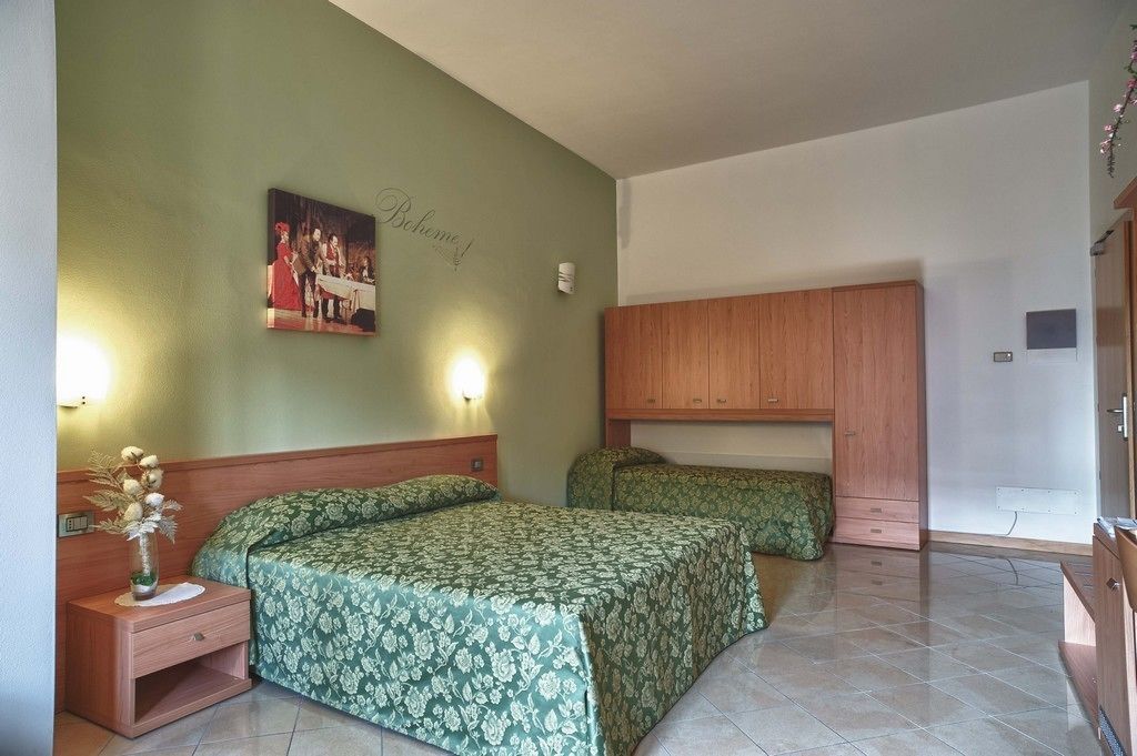 A room with two beds, a wooden wardrobe, a nightstand with a vase, and a wall-mounted light fixture.