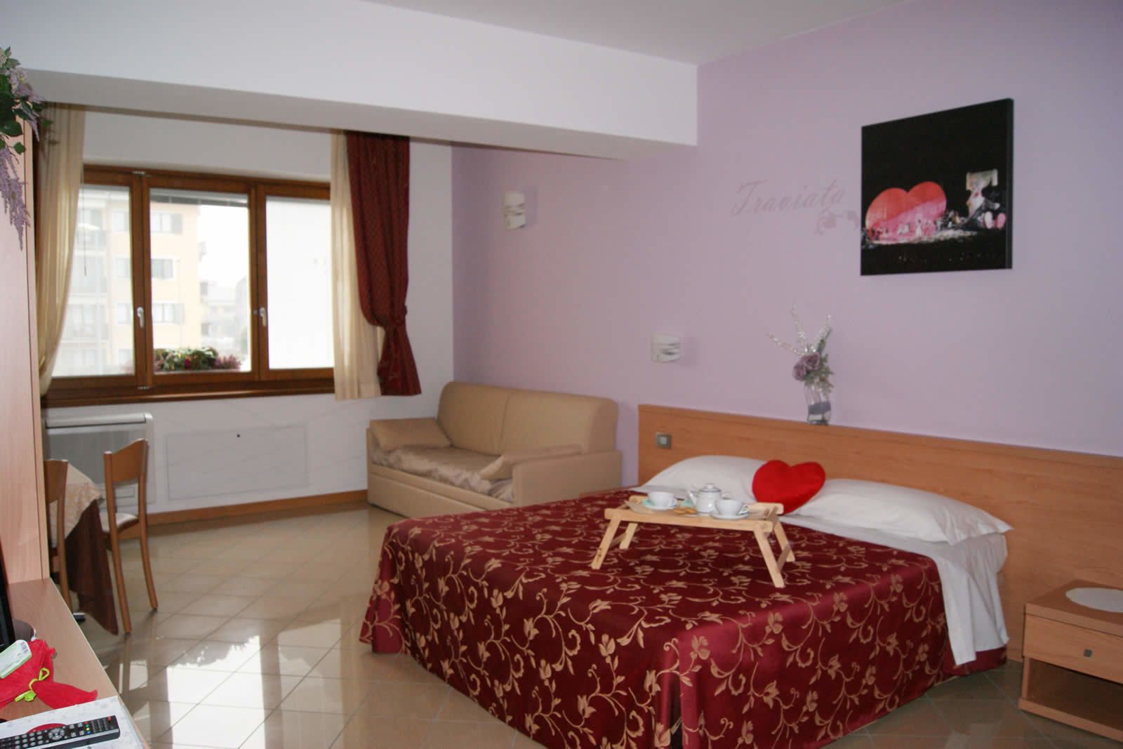 A hotel room with a bed, a chair, a small table with a tray, a window with curtains, and a wall with a picture.