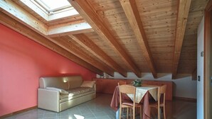 Un acogedor dormitorio en el ático con techo de madera, pared roja, sofá, mesa de comedor con sillas y un tragaluz.