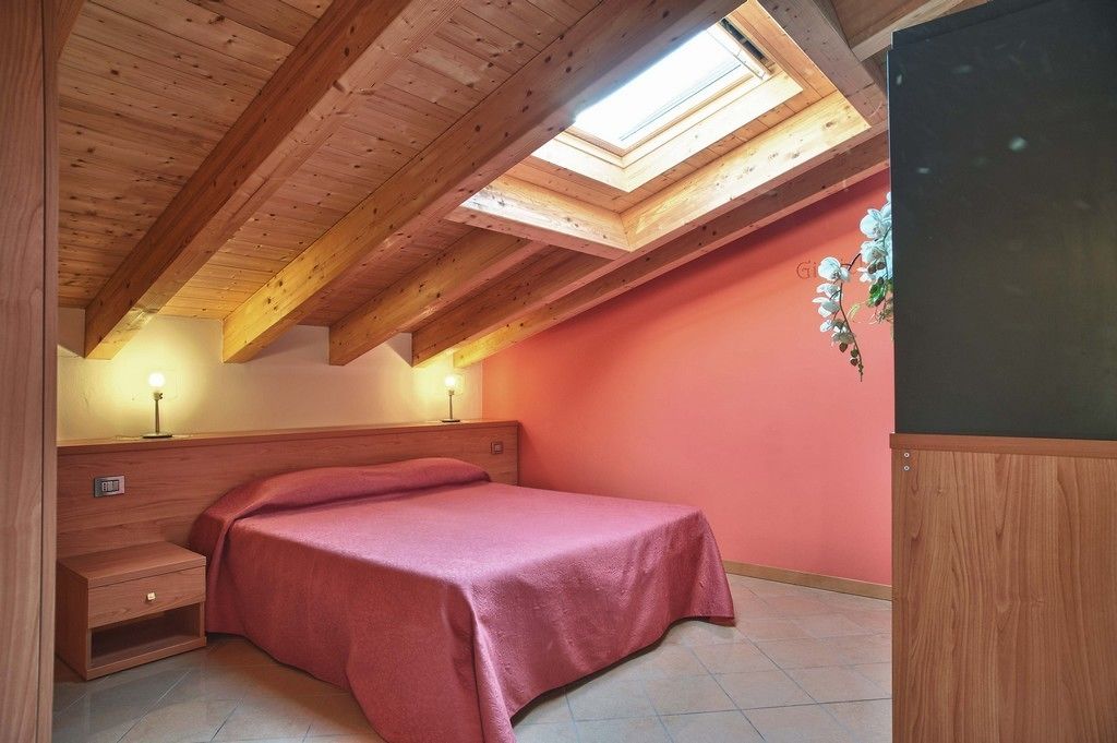 Een slaapkamer met een schuine kap, dakraam, een bed met een rode sprei, nachtkastje en een aan de muur bevestigde televisie.