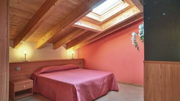 Een slaapkamer met een schuine kap, dakraam, een bed met een rode sprei, nachtkastje en een aan de muur bevestigde televisie.