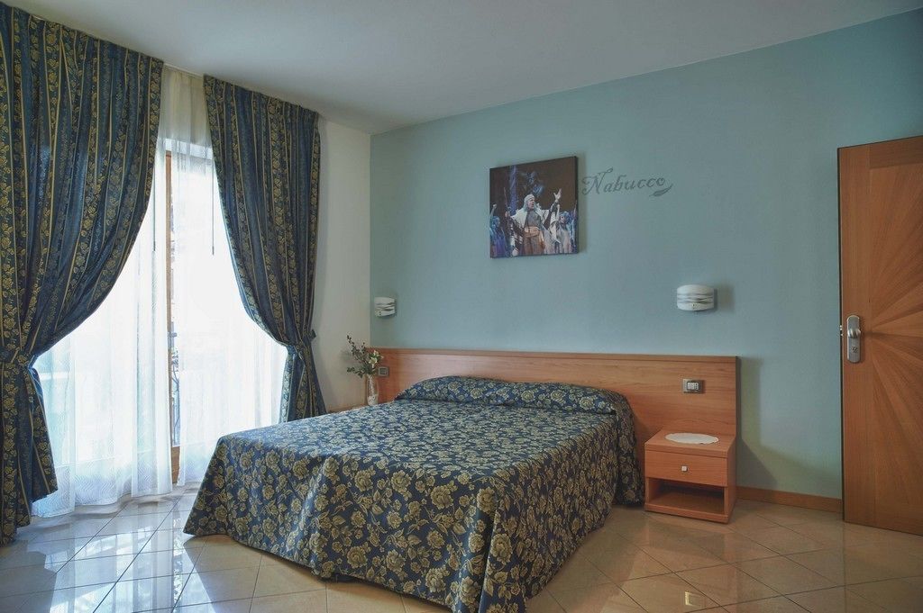 Une chambre à coucher comprenant un lit, une table de chevet, une lampe fixée au mur, une porte et une fenêtre avec des rideaux.