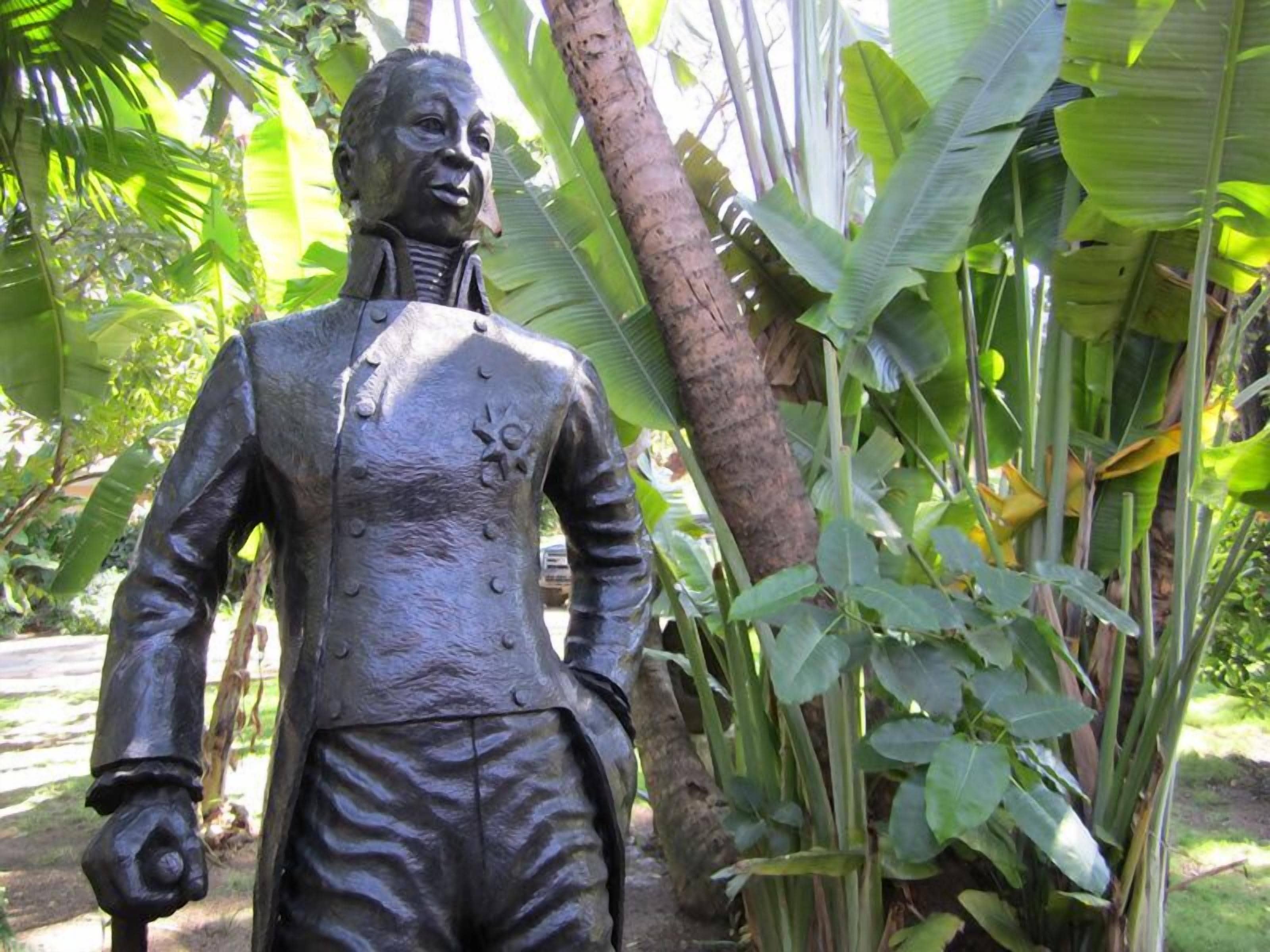 A statue of a man in a suit with a flower on his chest, surrounded by lush green foliage.