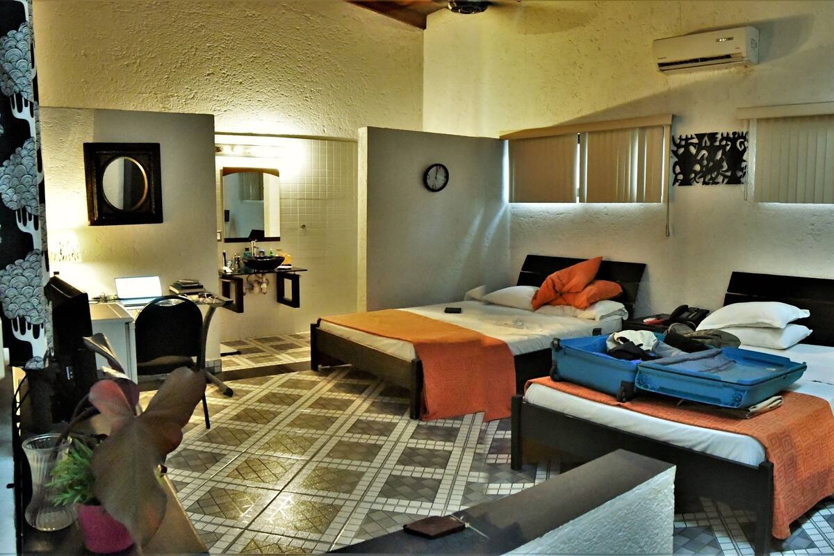 A compact hotel room with a bed, a desk, a chair, a small table, a mirror, a clock, and a window with blinds.