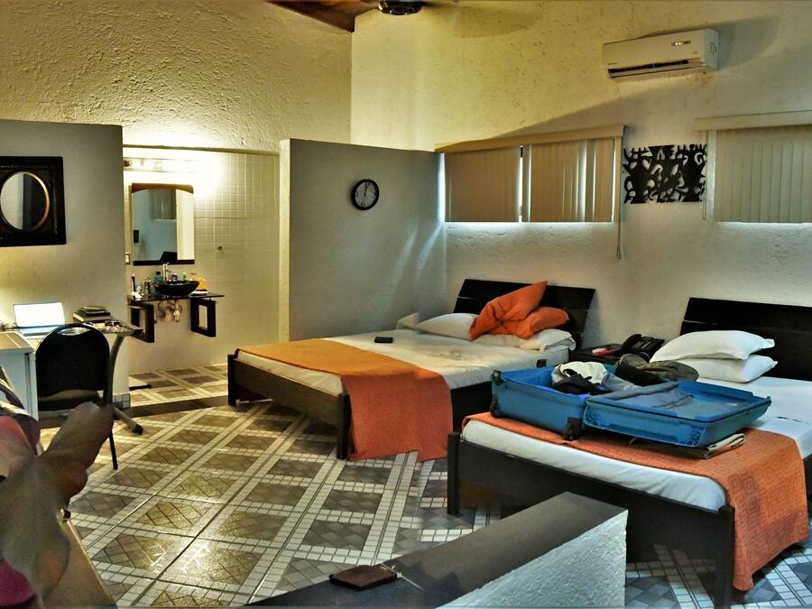 A compact hotel room with a bed, a desk, a chair, a small table, a mirror, a clock, and a window with blinds.