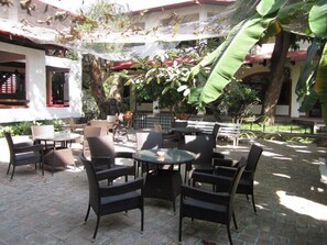 Un espace extérieur aménagé en terrasse, avec des meubles en osier, notamment des tables et des chaises rondes, entouré de plantes et d’arbres en pot.