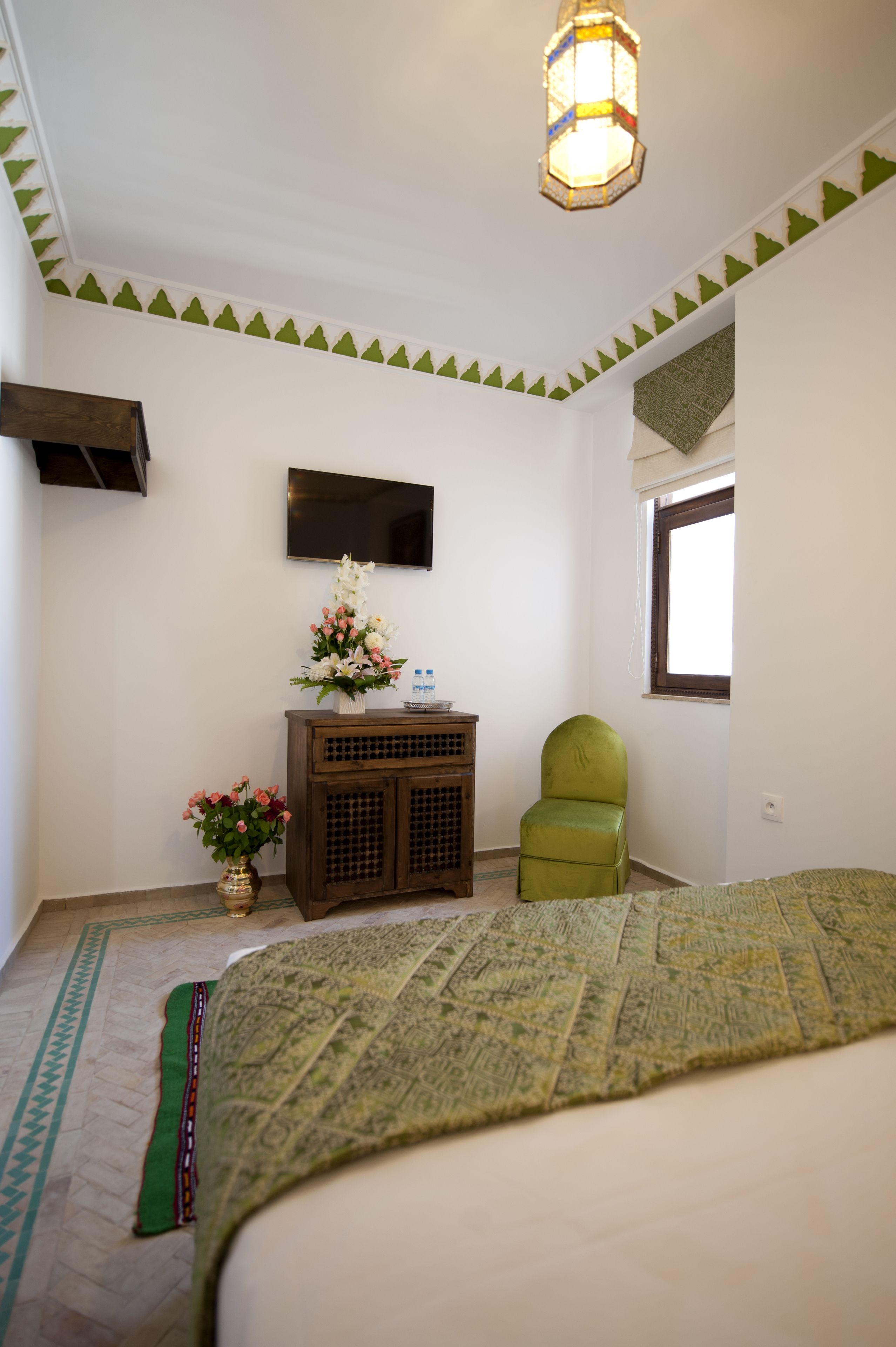 Un dormitorio con una cama, una silla verde, un armario de madera con un florero y un televisor de pantalla plana en la pared, además de una ventana con vistas.