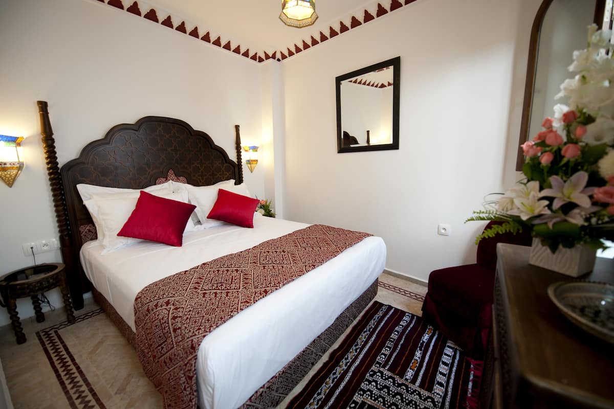 Una cama bien hecha con ropa de cama estampada y almohadas rojas, una mesita de noche pequeña, un espejo y un jarrón con flores.