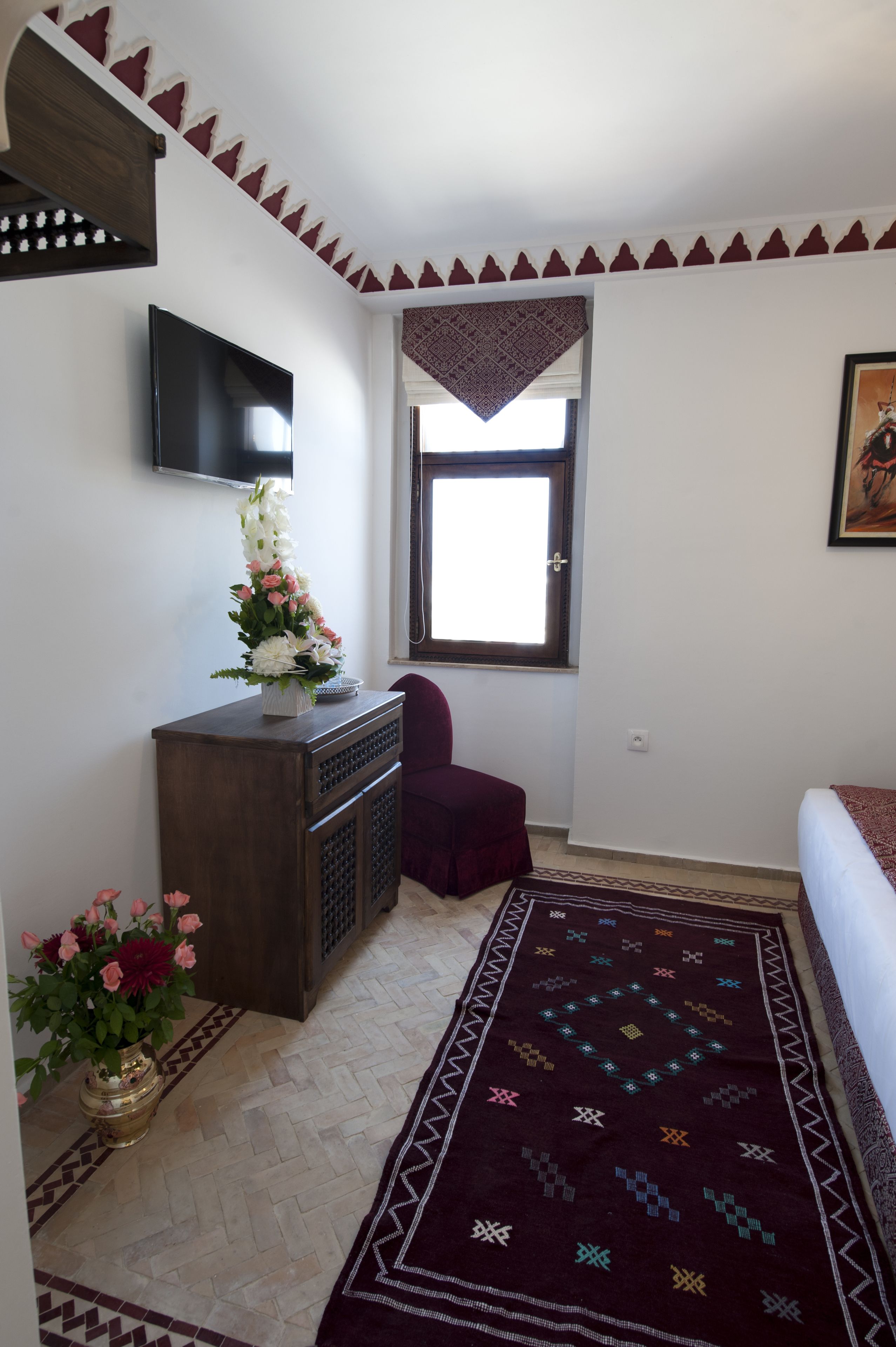A bedroom with a bed, a TV mounted on the wall, a window with a view, a decorative rug, and a small table with flowers.