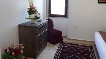 Un dormitorio con cama, televisor montado en la pared, ventana con vistas, alfombra decorativa y una mesita con flores.