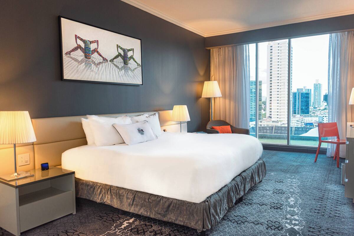 A hotel room with a large bed, two bedside tables, a desk, a chair, and a large window with a city view.