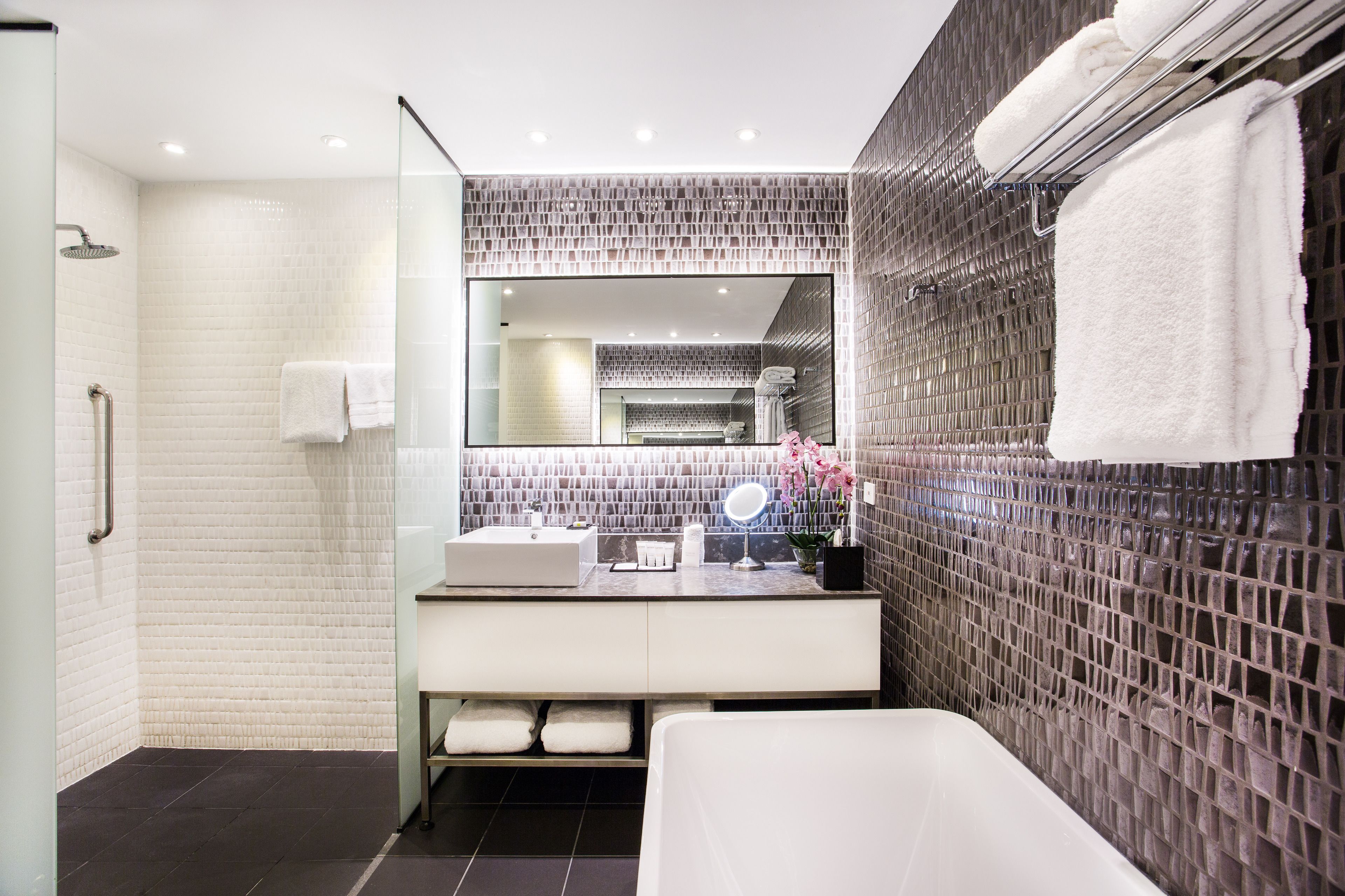 Un baño moderno con cabina de ducha de vidrio, un lavabo blanco con espejo y una pared revestida de baldosas oscuras.