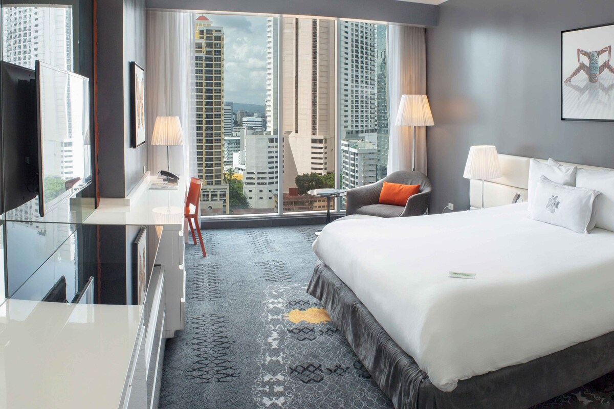 A hotel room with a large bed, two bedside tables, a desk with a chair, a TV, and a city view through large windows.