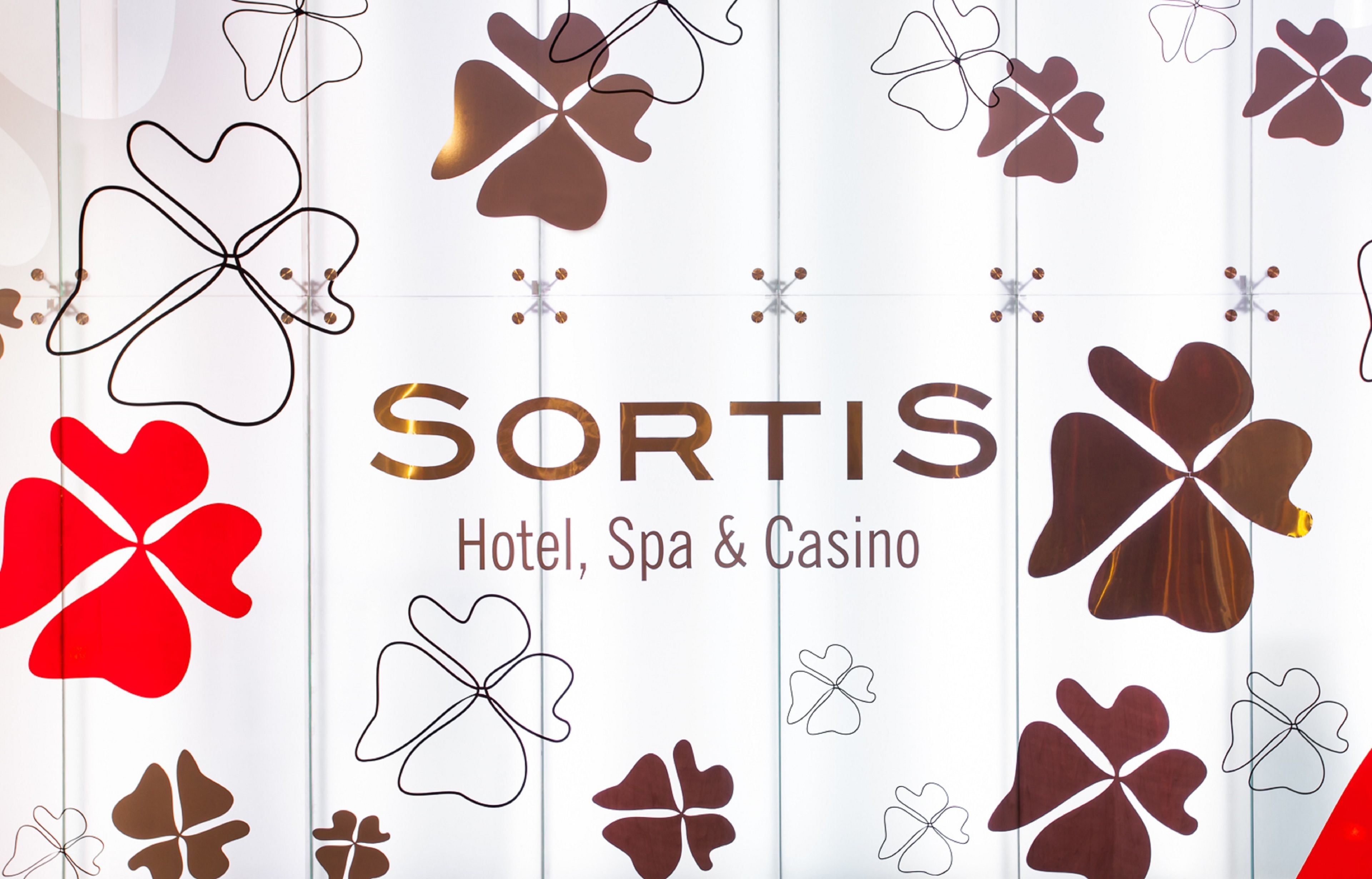 A promotional graphic for SORTIS Hotel, Spa & Casino with a pattern of stylized clover shapes and a gold text logo.