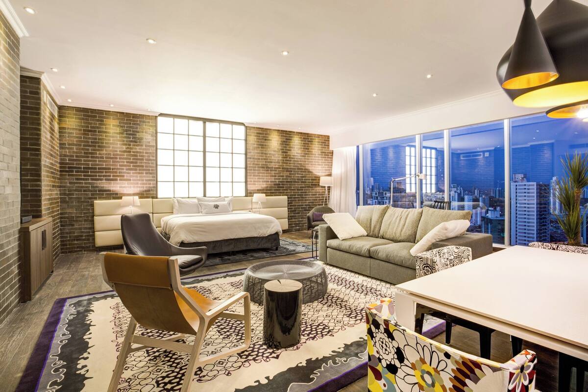 A modern hotel room with a large bed, a sofa, a dining table, and a city view through large windows.
