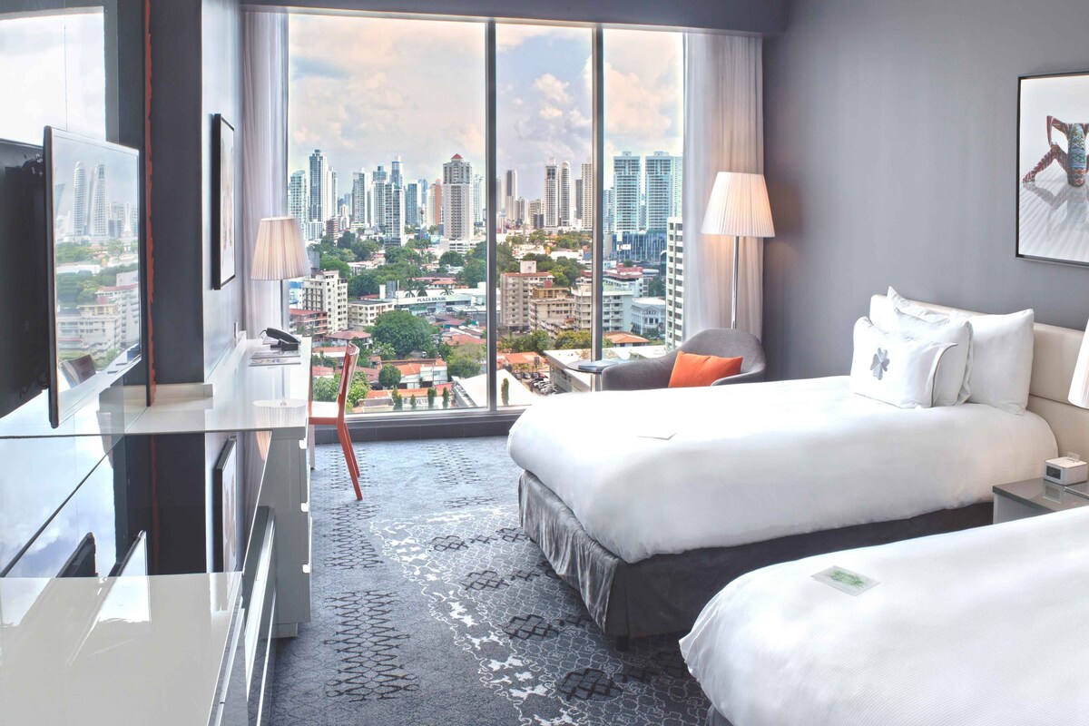 A hotel room with two beds, a desk, a chair, a TV, and a large window offering a city view.