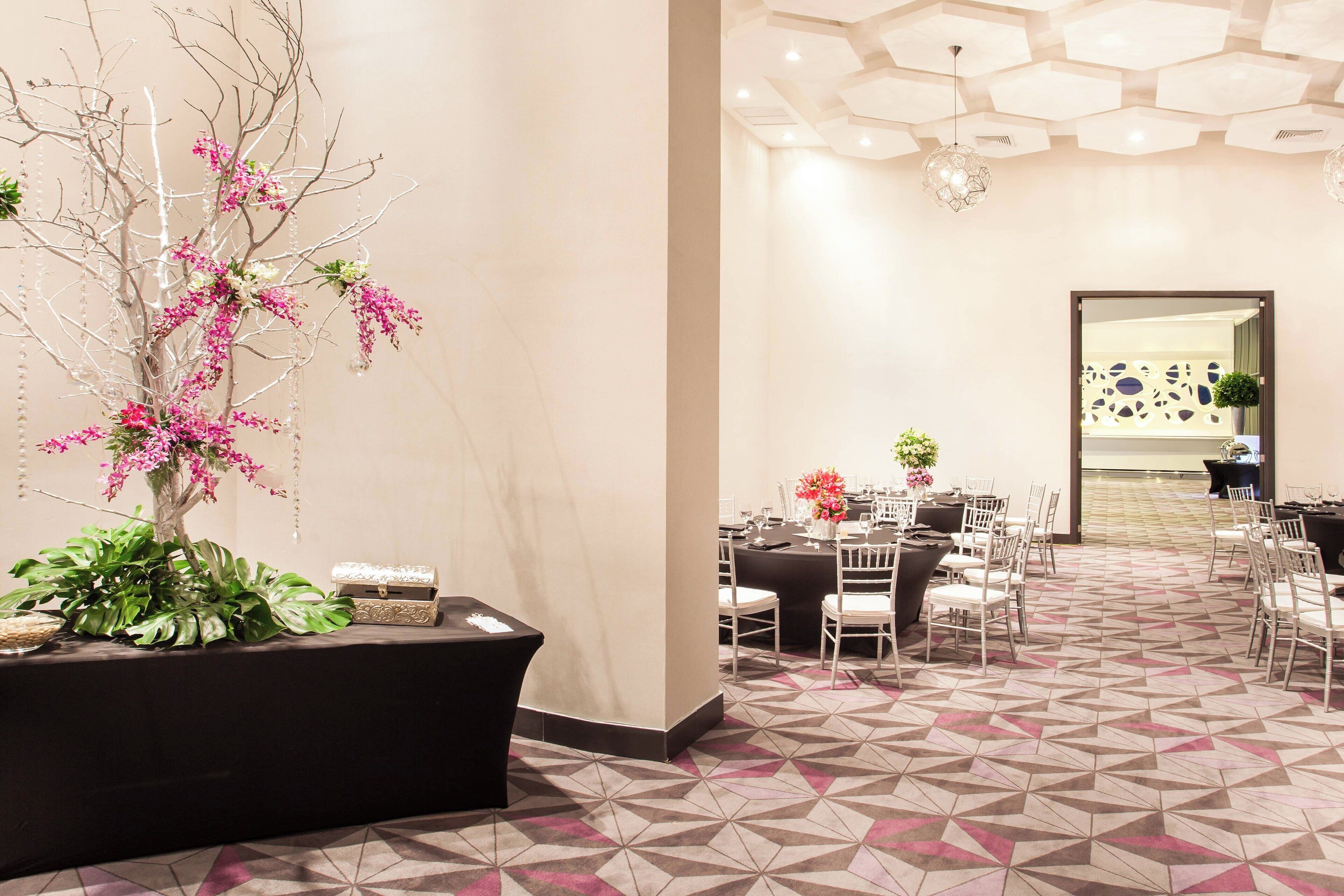 A hotel lobby with a geometric patterned floor, a large potted plant with pink flowers, and a seating area with tables and chairs.