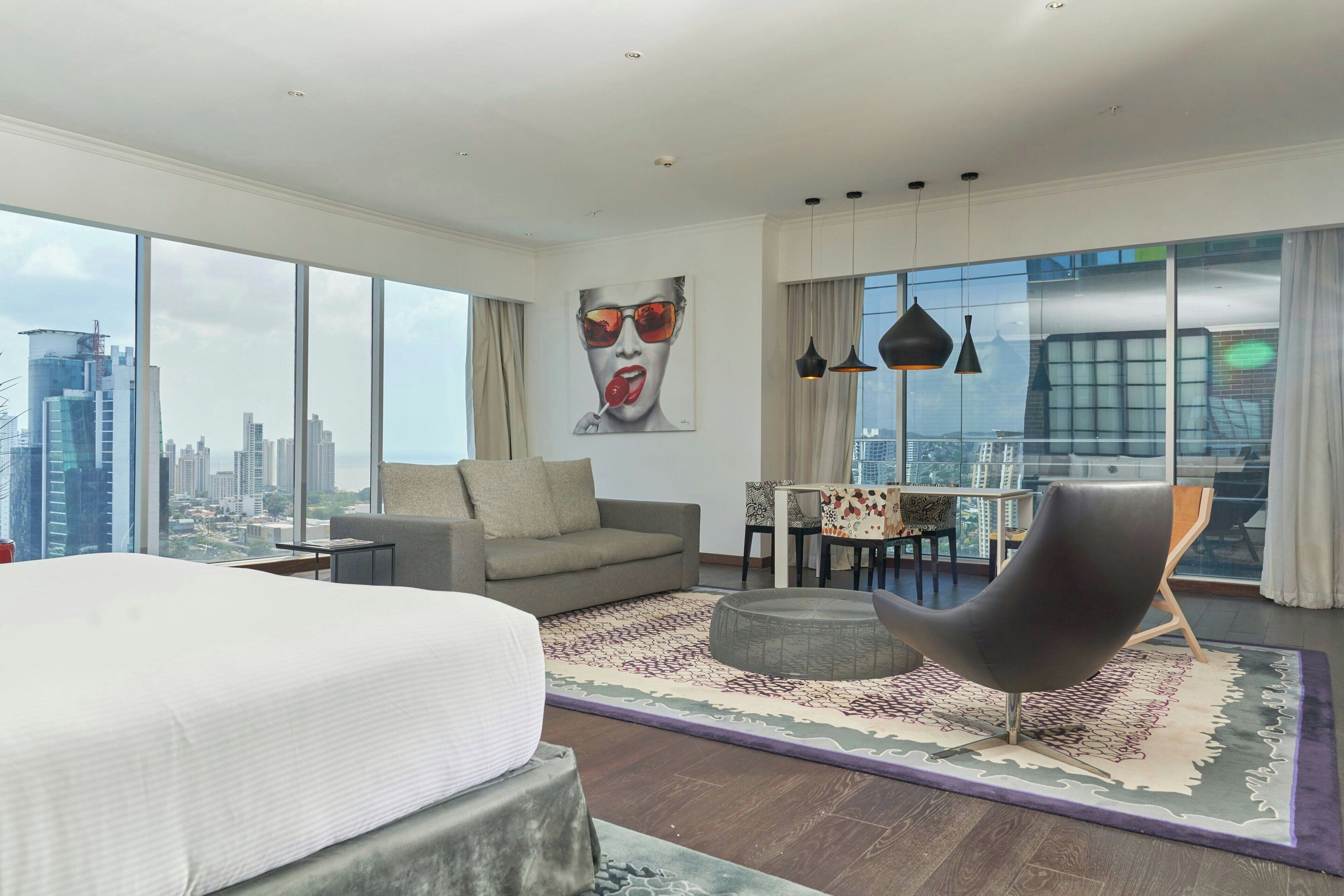 Una habitación de hotel moderna con una cama grande, un sofá gris, un sillón negro, una mesa de comedor con sillas y un amplio ventanal con vistas a la ciudad.