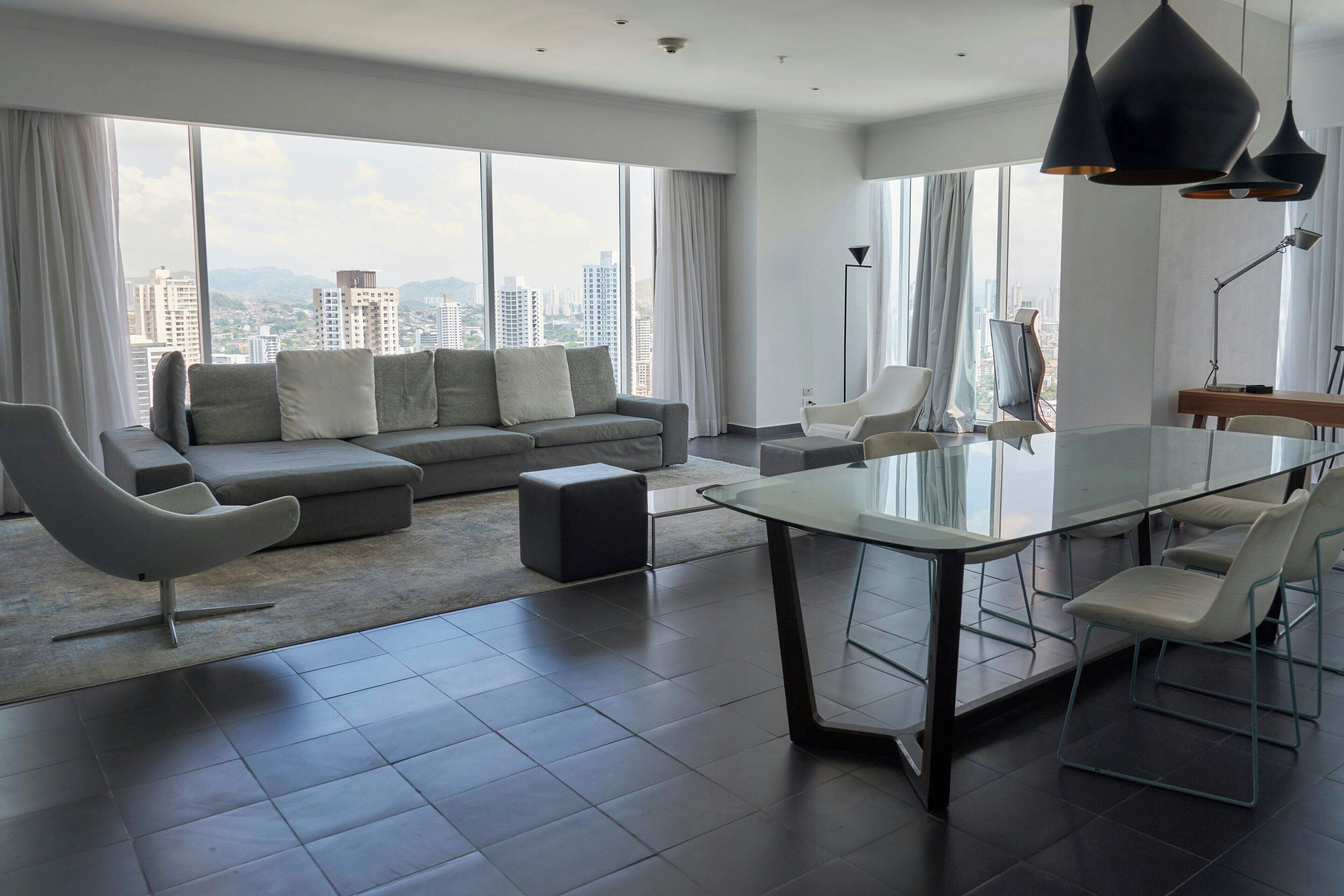A modern living room with a grey sofa, a glass dining table, and a city view.