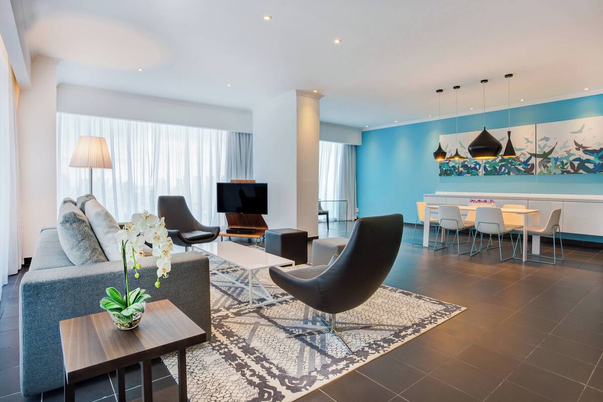 A modern living room with a sofa, armchairs, a coffee table, and a TV. There is a dining area with a table and chairs. The room has large windows with curtains, a blue accent wall, and a patterned rug.